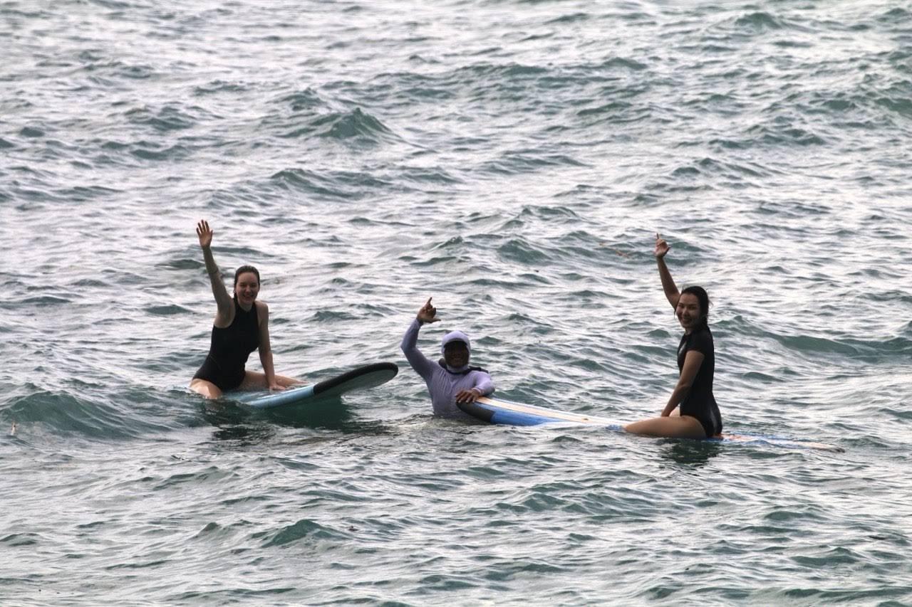 Arfas and two students waving from their surfboards in the ocean